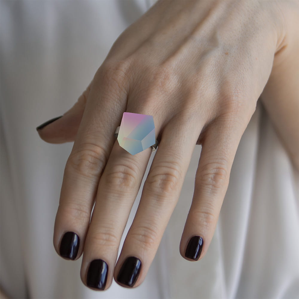 A hand with dark purple nail polish wears the Fruit Bijoux Ring Top Unicorn by Fruit Bijoux, featuring a geometric pastel gemstone set in sterling silver against a soft white fabric background.