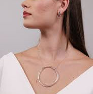 A woman with long dark hair is shown in profile wearing a delicate silver-toned necklace with a large circular pendant and small hoop earrings against a light gray background.