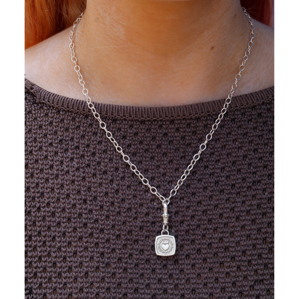 Worn over a brown textured sweater, The Collective Dublin’s Keith Haring Inspired Claddagh Pendant with chunky chain features a square pendant engraved with a heart and sunburst motif, echoing the style of an Irish classic necklace.