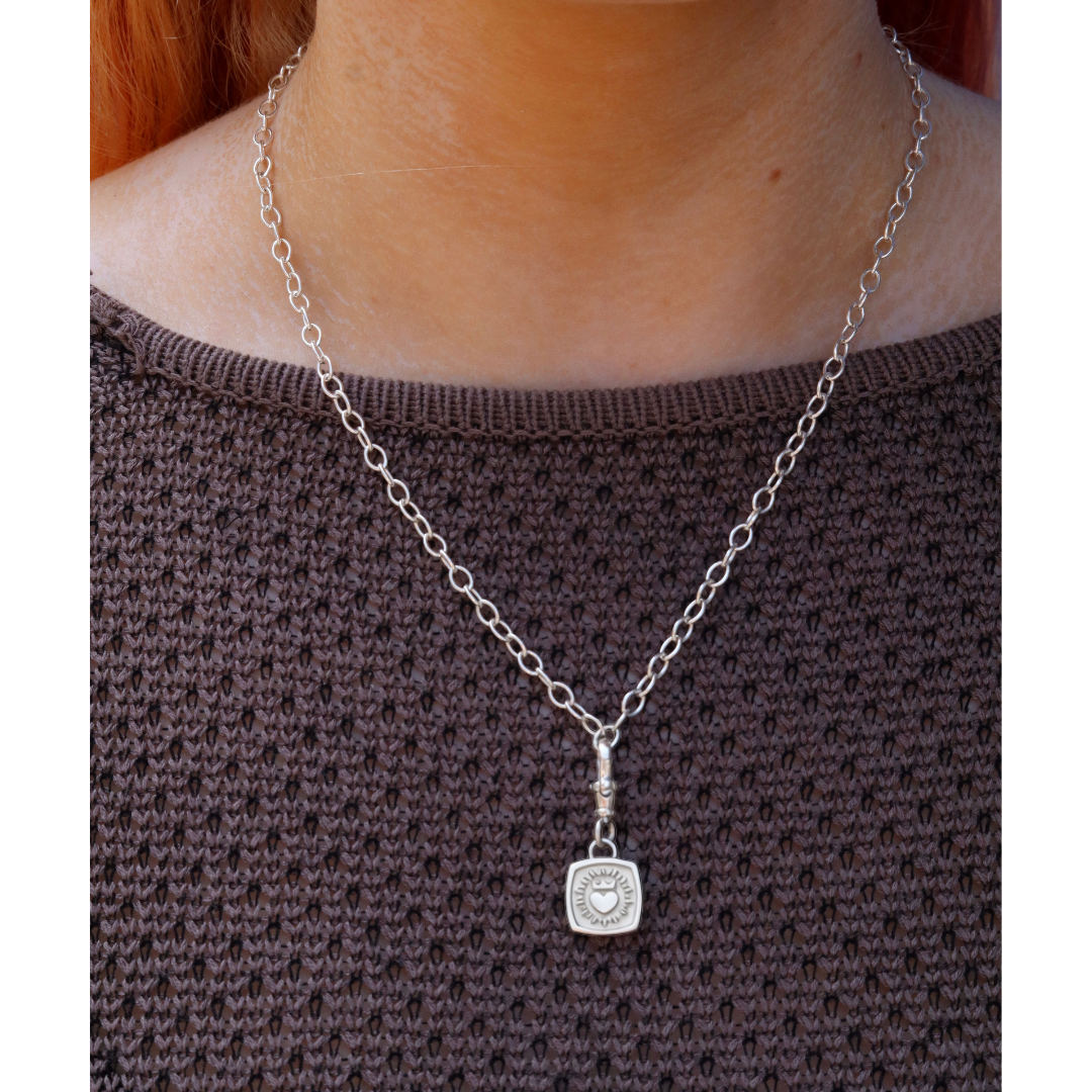 Worn over a brown textured sweater, The Collective Dublin’s Keith Haring Inspired Claddagh Pendant with chunky chain features a square pendant engraved with a heart and sunburst motif, echoing the style of an Irish classic necklace.