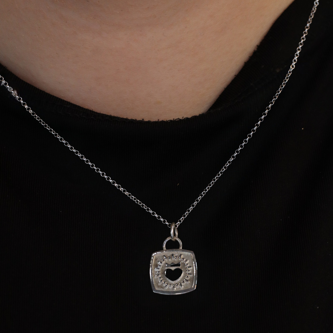 A Keith Haring Inspired Claddagh Pendant necklace by The Collective Dublin, featuring a square sterling silver pendant with a heart-shaped cutout, is worn on the neck over a black shirt. This piece brings a subtle touch of Irish jewelry elegance to any outfit.