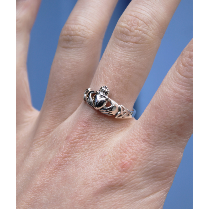 A close-up of a hand wearing the Watermelon tropical Claddagh Ring Sterling Silver, featuring a crowned heart held by two hands—symbolizing love and friendship—set against a blue background.