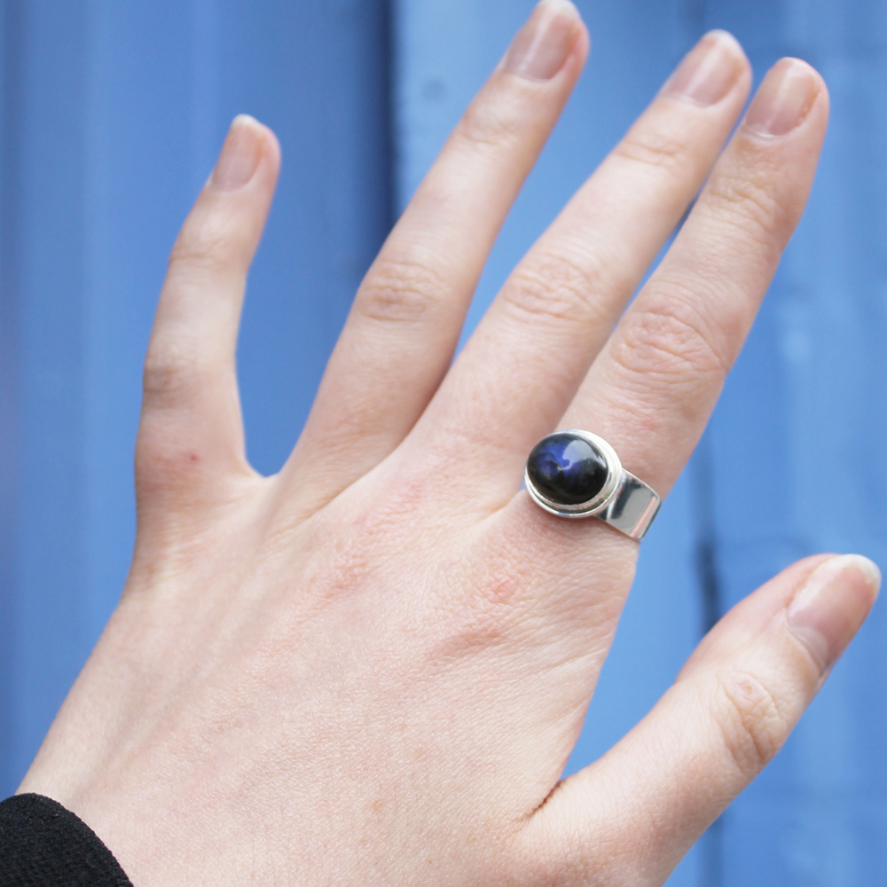 A hand displays the Watermelon tropical Horizontal Labradorite Sterling Silver Ring, featuring a dark blue labradorite stone, against a blue background.