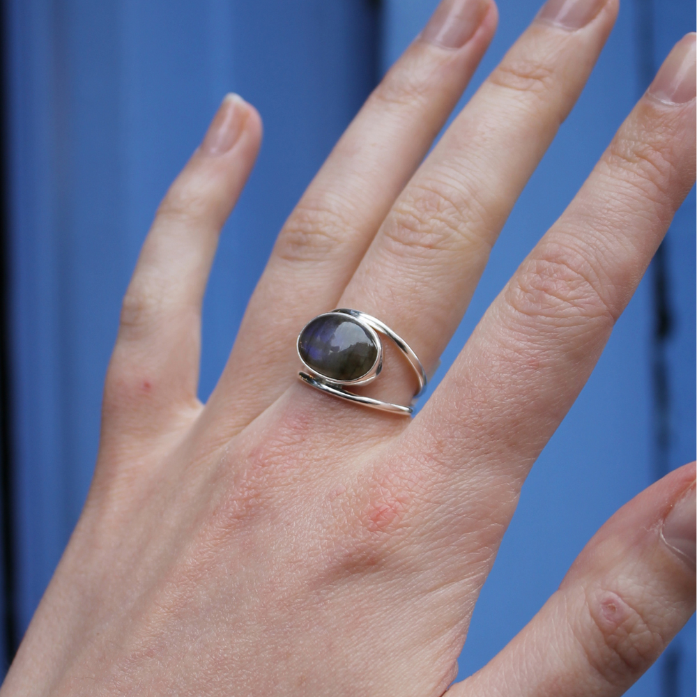 A hand wears the Watermelon tropical Double Silver Band Labradorite Ring, featuring an oval dark iridescent stone, on the middle finger against a blue background. The nails are natural and unpolished.