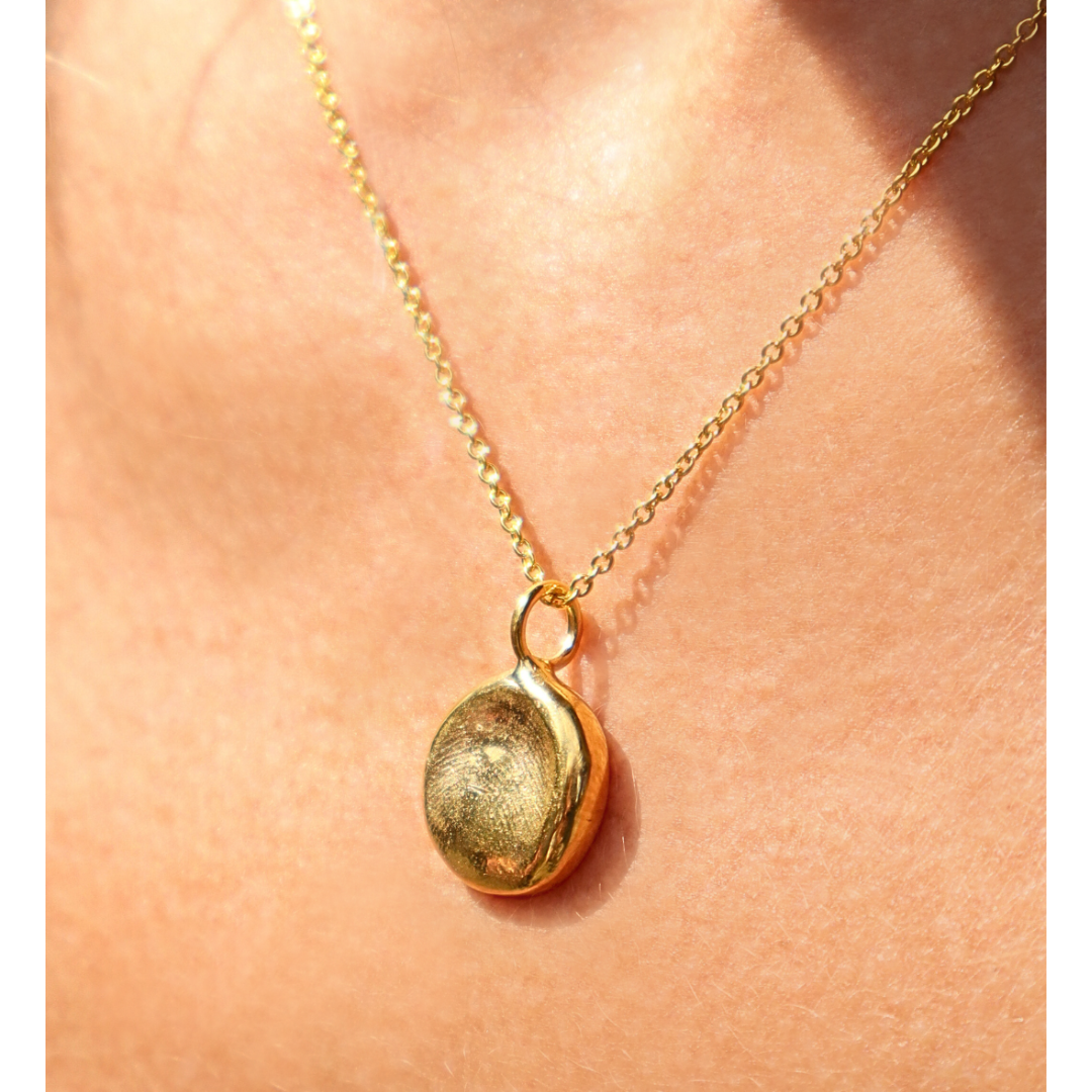 The Silver Works Fingerprint Imprint Necklace features a gold pendant with a round, textured charm, worn close to the skin in sunlight—perfect as a personalized memorial keepsake or unique gift.