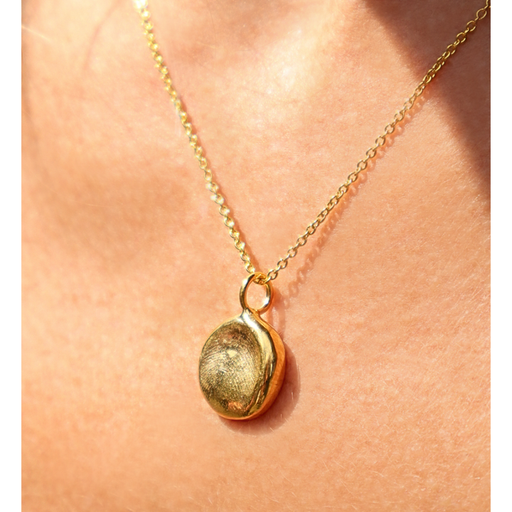 The Silver Works Fingerprint Imprint Necklace features a gold pendant with a round, textured charm, worn close to the skin in sunlight—perfect as a personalized memorial keepsake or unique gift.