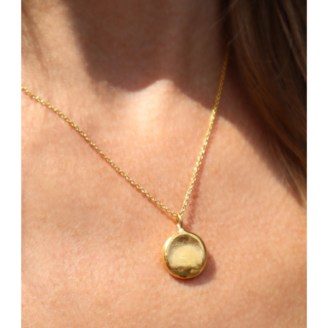 A close-up of a person wearing Silver Works’ Fingerprint Imprint Necklace, a gold chain with a round, smooth memorial keepsake pendant, shown against their bare skin with partially visible hair.