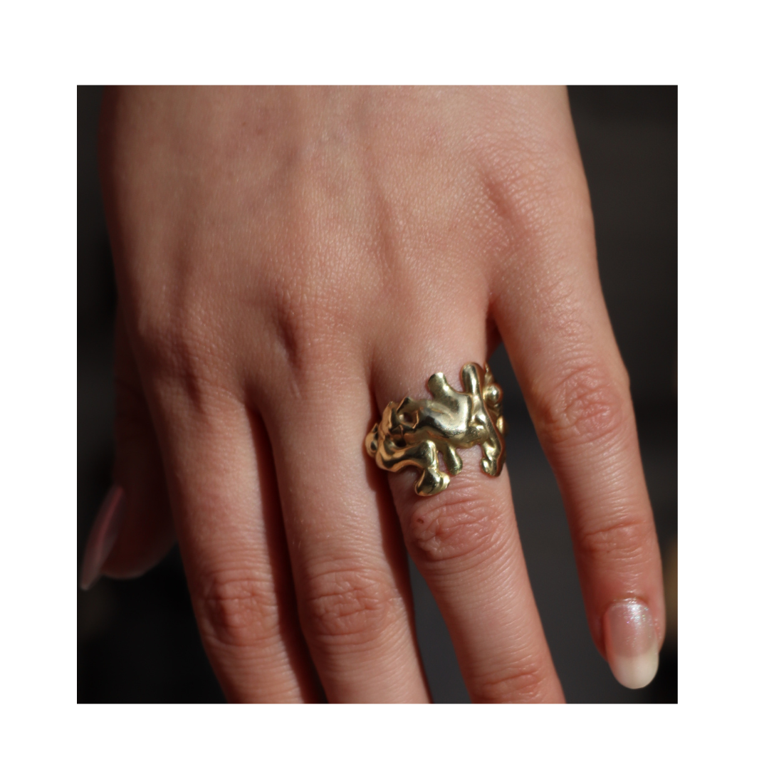 A close-up of a hand wearing the Molten Ring by Madeleine Sculptural Jewellery on the middle finger. The chunky, abstract gold piece shines against natural nails, set against a dark, blurred background.