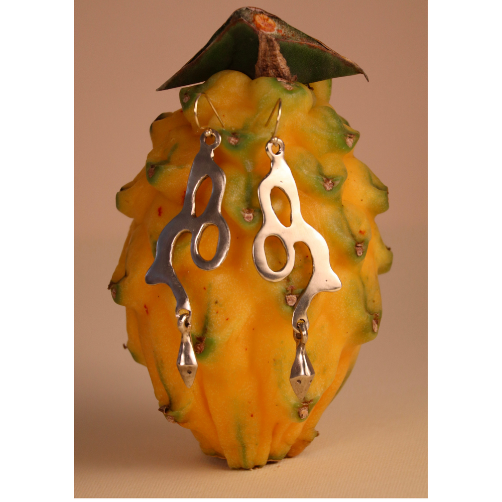 The Serpent Earrings by Madeleine Sculptural Jewellery, featuring an abstract silver design, hang from a yellow dragon fruit with green-tipped scales, set against a beige background.