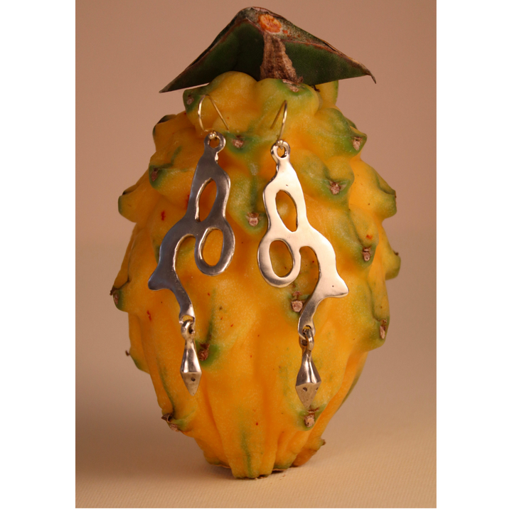 The Serpent Earrings by Madeleine Sculptural Jewellery, featuring an abstract silver design, hang from a yellow dragon fruit with green-tipped scales, set against a beige background.