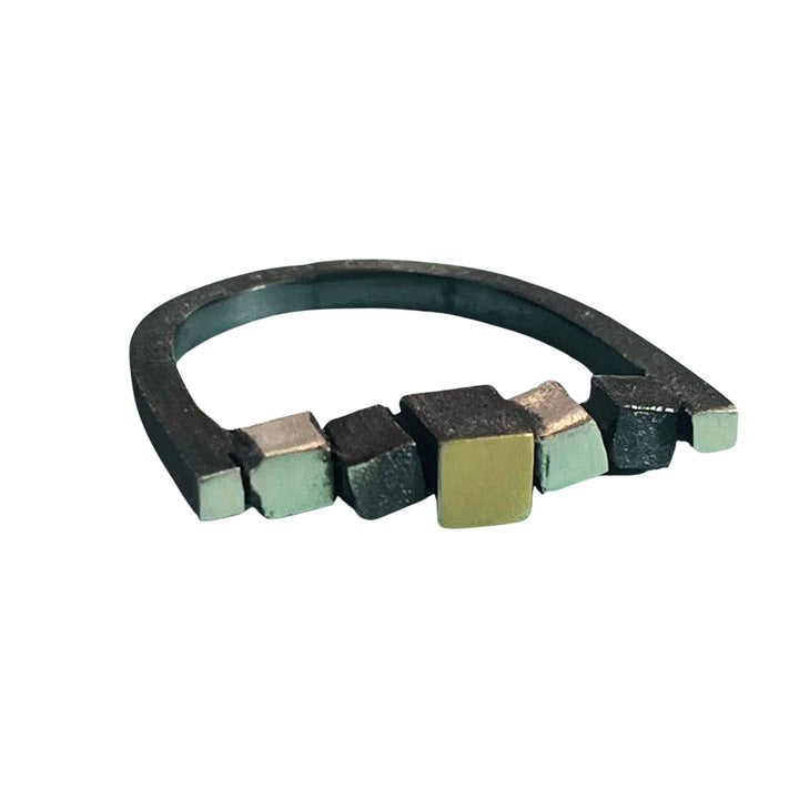 A unique, contemporary ring from the Iceland collection featuring a blackened metal band with various small, geometric cubes in different metallic shades attached along the top. The cubes are in matt silver, black, and 18kt yellow gold tones, creating a modern, artistic look. Introducing the MarCucurella Narrow Iceland Ring.