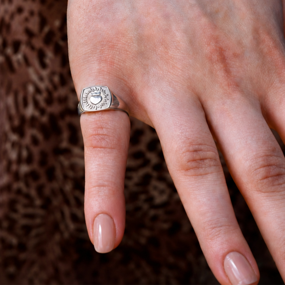 A close-up of a light-skinned hand wearing The Collective Dublin’s handmade Keith Haring Inspired Pinky Claddagh Ring, nails neatly manicured, with a brown patterned background.