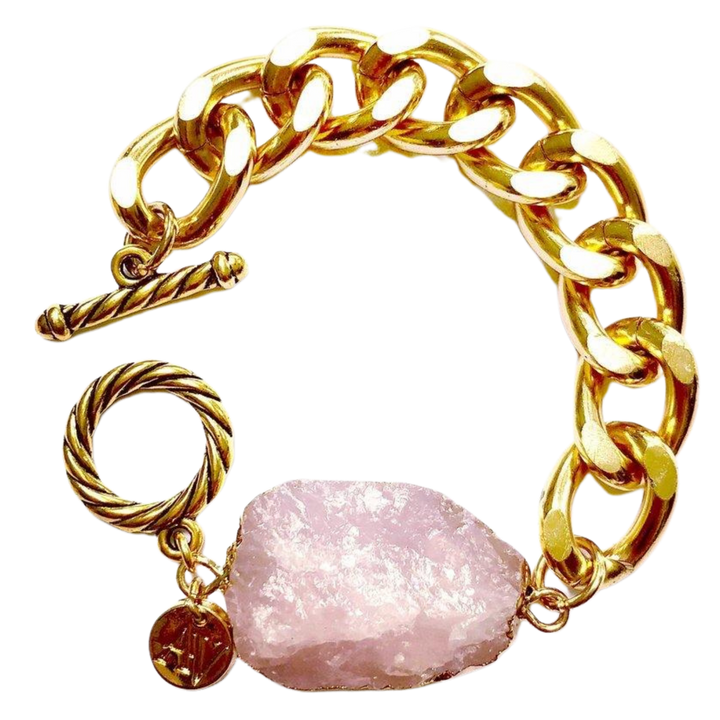 The Cruise Chunky Bracelet by Aria V features a large, rough rose quartz stone centerpiece. The bracelet has a toggle clasp with rope-like detailing and a small circular charm attached. Handmade in Dublin, the chain links are thick and polished.