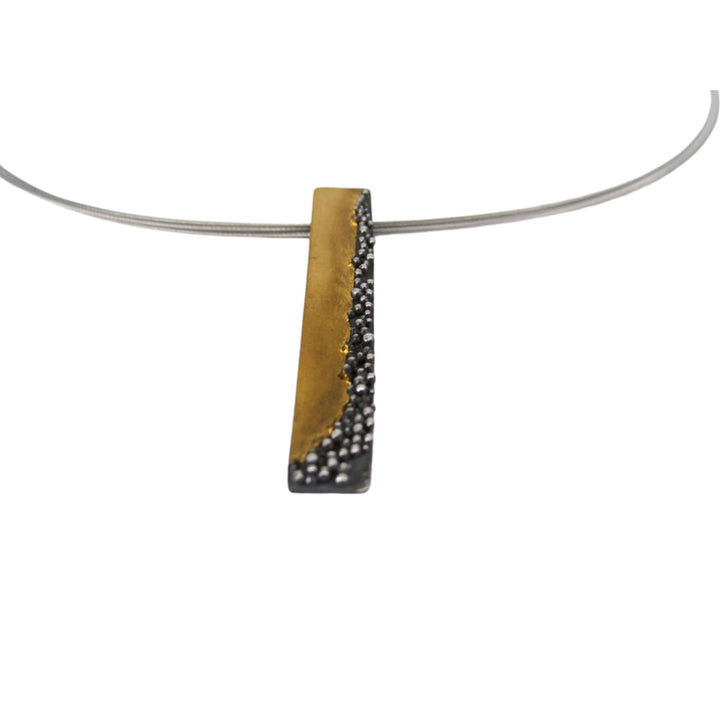 A minimalist rectangular pendant hangs on a thin, silver wire necklace. The Etrusc Pendant by MarCucurella features a two-tone design with a smooth section of 24 kt yellow gold on the left and a textured, oxidized silver section with small silver dots on the right.