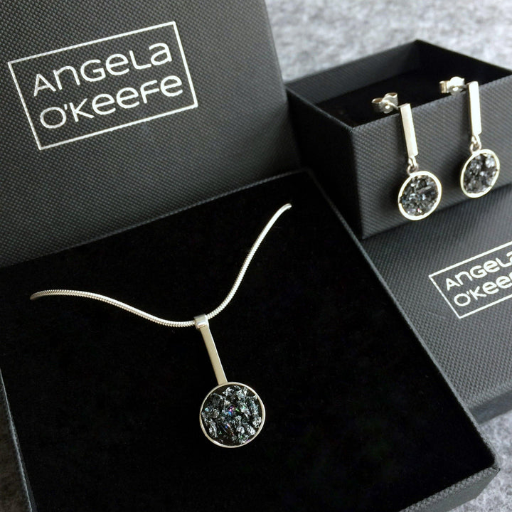 An Urban Geode Silver Dome Drop Pendant displayed in an Angela O'Keefe branded box. The set includes a silver pendant necklace and a pair of earrings. Each piece features a circular, textured, silver-colored design. The box interior is lined with black material, providing contrast to the shiny jewelry, evoking a gothic fairy tale charm.