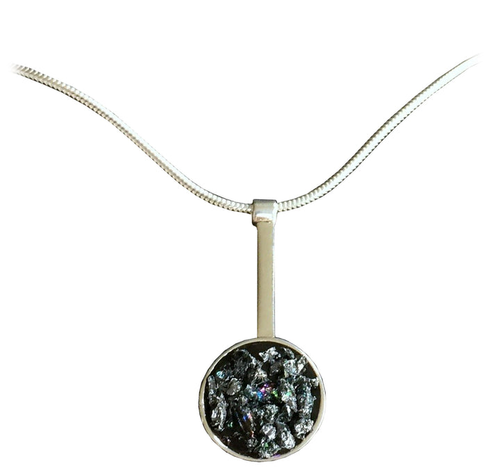 A silver necklace featuring a pendant with a round, rough-textured dark mineral encased in a slim, vertical metallic holder. The necklace has a fine, smooth chain, creating a simple yet elegant look reminiscent of a gothic fairy tale. The background is plain white. Introducing the Urban Geode Silver Dome Drop Pendant by Angela O'Keefe.