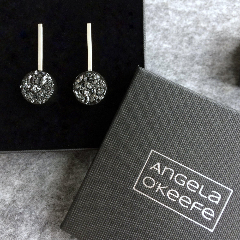 A pair of Urban Geode Silver Sphere Drop Earrings from the Angela O'Keefe collection is displayed inside a black box. Beside the box is another smaller, closed black box with the name "Angela O'Keefe" branded on the lid in silver text.