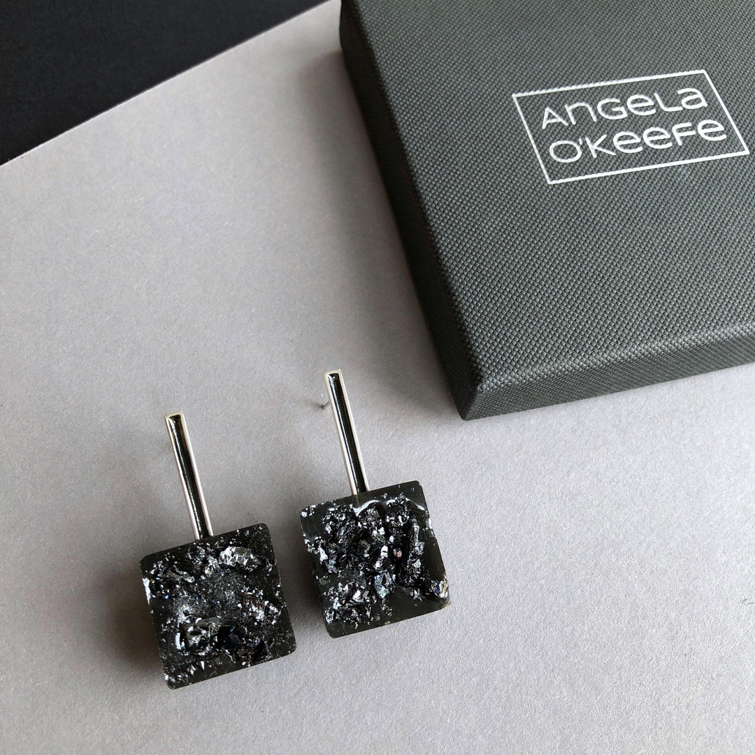 A pair of Angela O'Keefe Urban Geode Square Black Resin Earrings with black textured square stones and sleek metal stems is shown on a gray and black surface. To the right, there is a dark gray box with the name "Angela O'Keefe" written on the lid in white text.