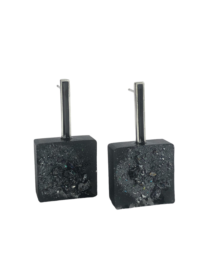 A pair of black geometric earrings with a rectangular post and square base. The surface of the square base has a textured design with sparkling, irregular surfaces that resemble crushed crystals. These Urban Geode Square Black Resin Earrings by Angela O’Keefe boast a modern and edgy appearance.