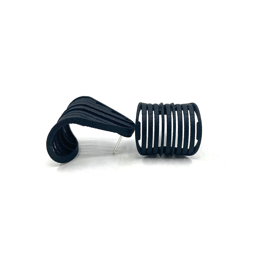 A pair of unique, black modernist earrings made from flat, segmented strips creating a wave-like structure. Crafted as Elsiem Vibe Grill Earrings with sterling silver ear posts, one earring is laid flat showing its backing pin, while the other stands upright, accentuating its spiraling design.