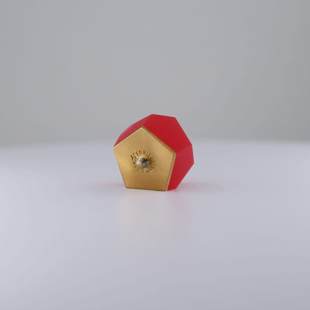 A Fruit Bijoux Ring Top VU Crystals Ruby Red from Fruit Bijoux with a red body and a 24ct gold-plated end lies on a smooth white surface against a light gray background.