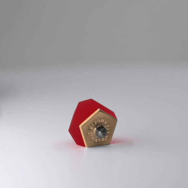The Fruit Bijoux Ring Top VU Crystals Ruby Red by Fruit Bijoux, featuring a ruby red pentagon design and brass base with an interchangeable ring module, rests on its side on a smooth white surface in soft, minimal lighting.