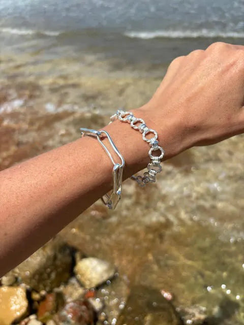 A hand adorned with two Shyla bracelets—including the Chunky Link Bracelet in silver—is extended over a rocky shore with waves, showcasing interlocking circular links and geometric patterns.