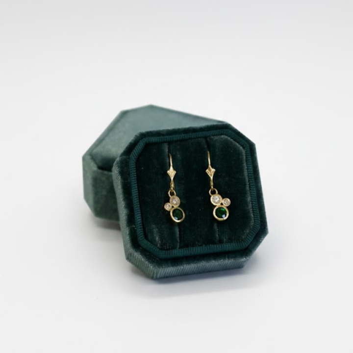 The Verdant Embrace 9ct yellow gold earrings by Madeleine Fine Jewellery, featuring green tsavorite and two diamonds, are displayed in a dark green velvet hexagonal jewelry box against a white background.