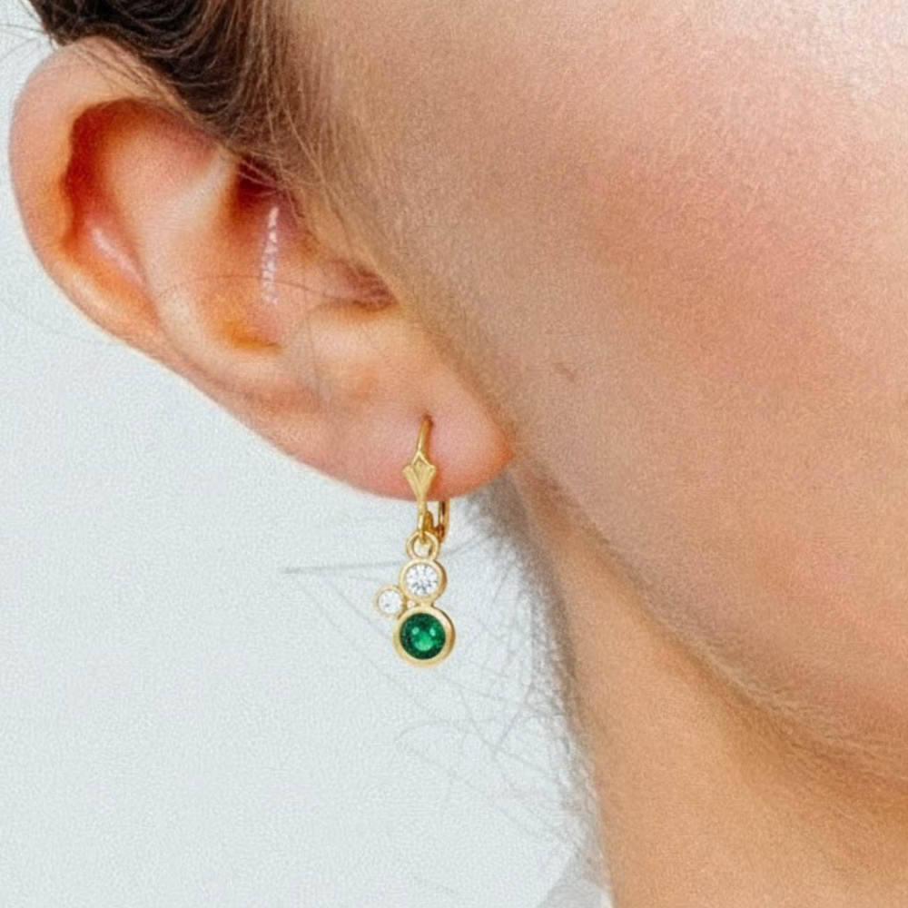 Close-up of a person's ear wearing Madeleine Fine Jewellery's Verdant Embrace 9ct yellow gold earrings, featuring green tsavorite and two diamonds. The background is plain and light-colored.