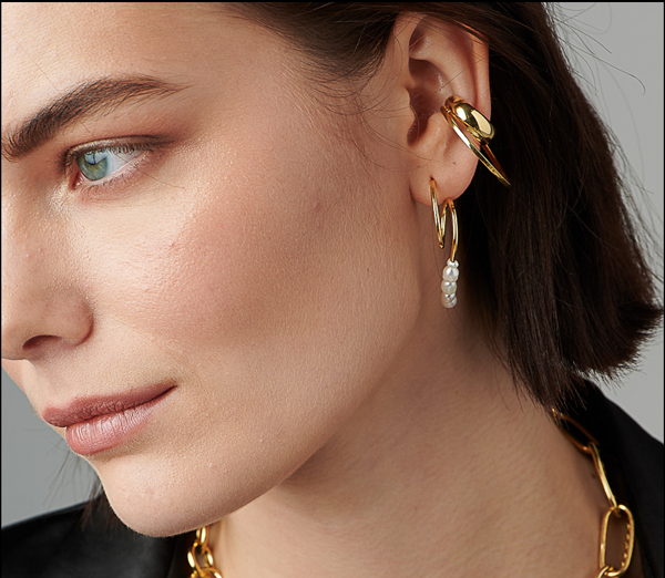 Close-up of a woman wearing gold jewelry. She has a gold chain necklace, a Bonvo Vero Ear Cuff with a heart motif, and a gold hoop earring adorned with small pearls. Her hair is short, and she is wearing minimal makeup. The background is neutral gray.