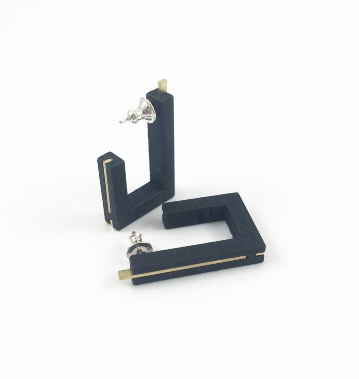 Vibe Brutalist Earrings by Elsiem feature a black geometric design with a minimalist aesthetic, shaped like the letter 'C' and reminiscent of Brutalist Architecture. One earring stands upright while the other lies flat, both displaying a silver stud attached to the top. The background is plain white.