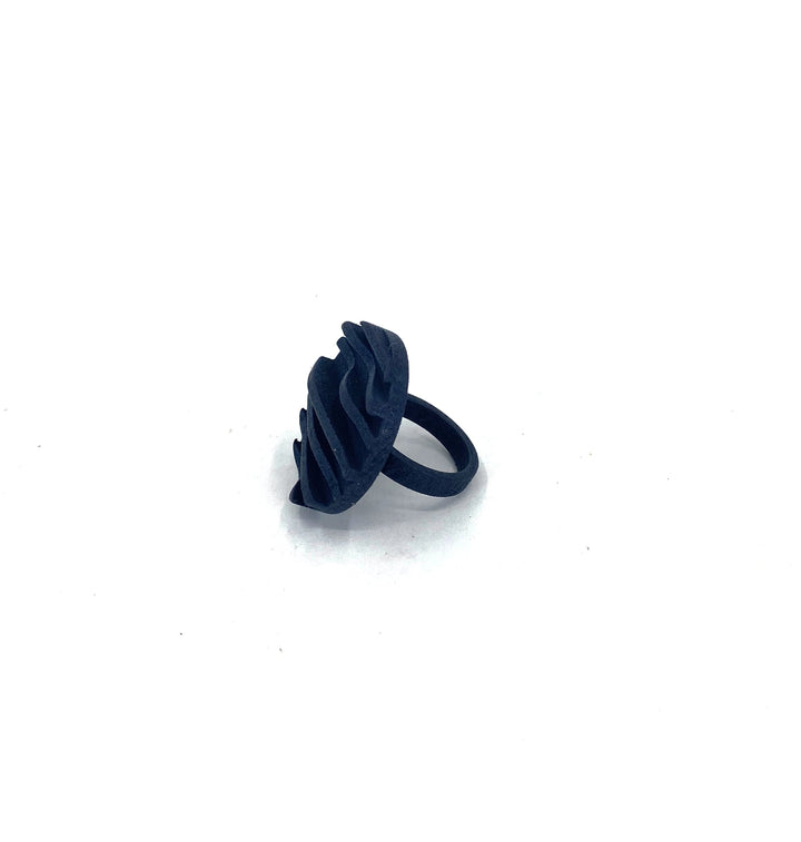 The Vibe Wave Circle Fascia Ring by Elsiem is a dark blue, 3D printed piece featuring a unique, layered, and wavy design on the top. Positioned upright against a plain white background, it showcases its artistic and abstract style as modern jewelry.