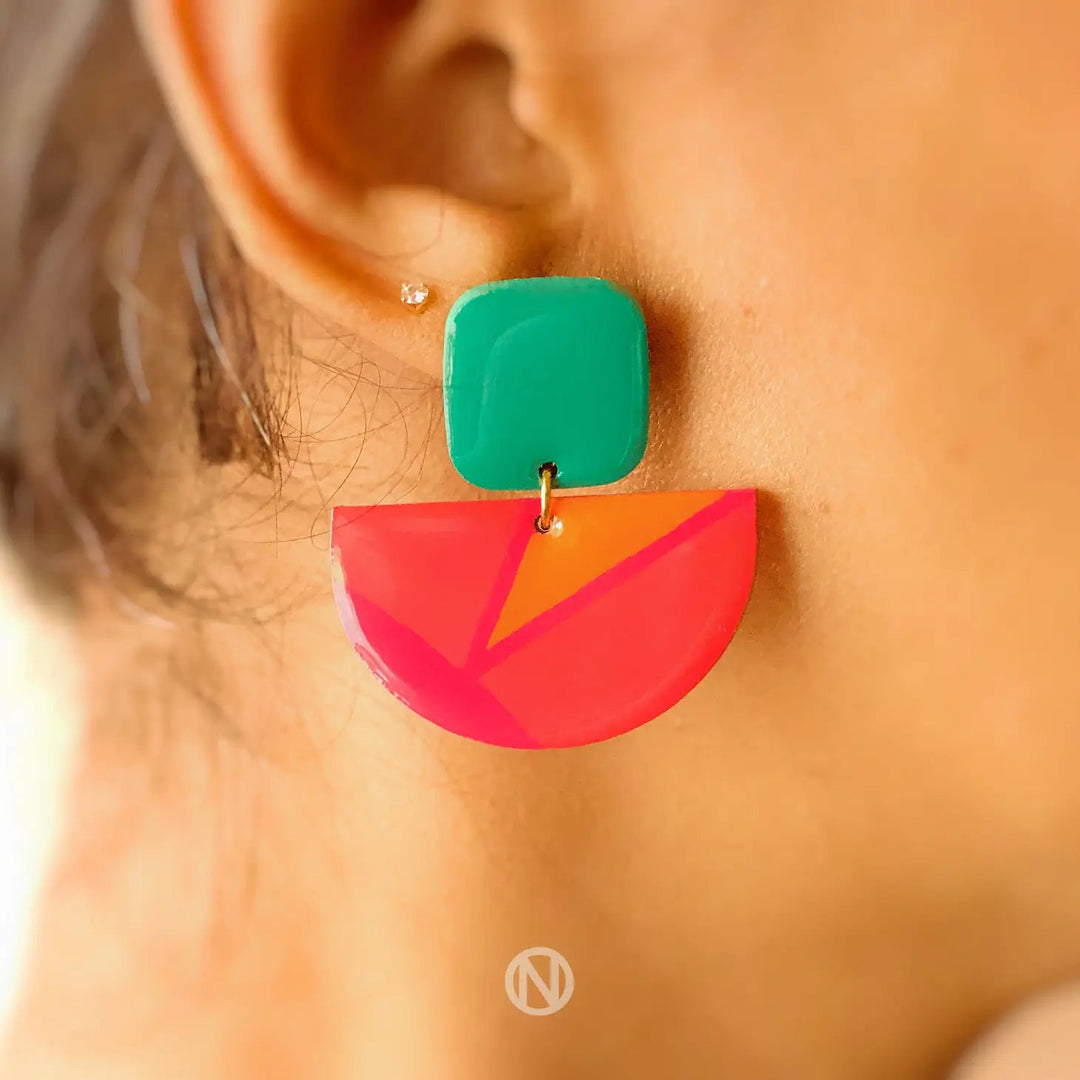 Close-up of a person's ear wearing Naoi Vivid Statement Earrings featuring a green square stud connected to a pink and orange hand-painted semi-circular pendant. A small, separate stud is visible higher on the ear. A logo is seen at the bottom center.