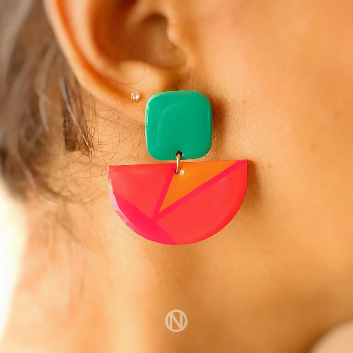 Close-up of a person's ear wearing Naoi Vivid Statement Earrings featuring a green square stud connected to a pink and orange hand-painted semi-circular pendant. A small, separate stud is visible higher on the ear. A logo is seen at the bottom center.