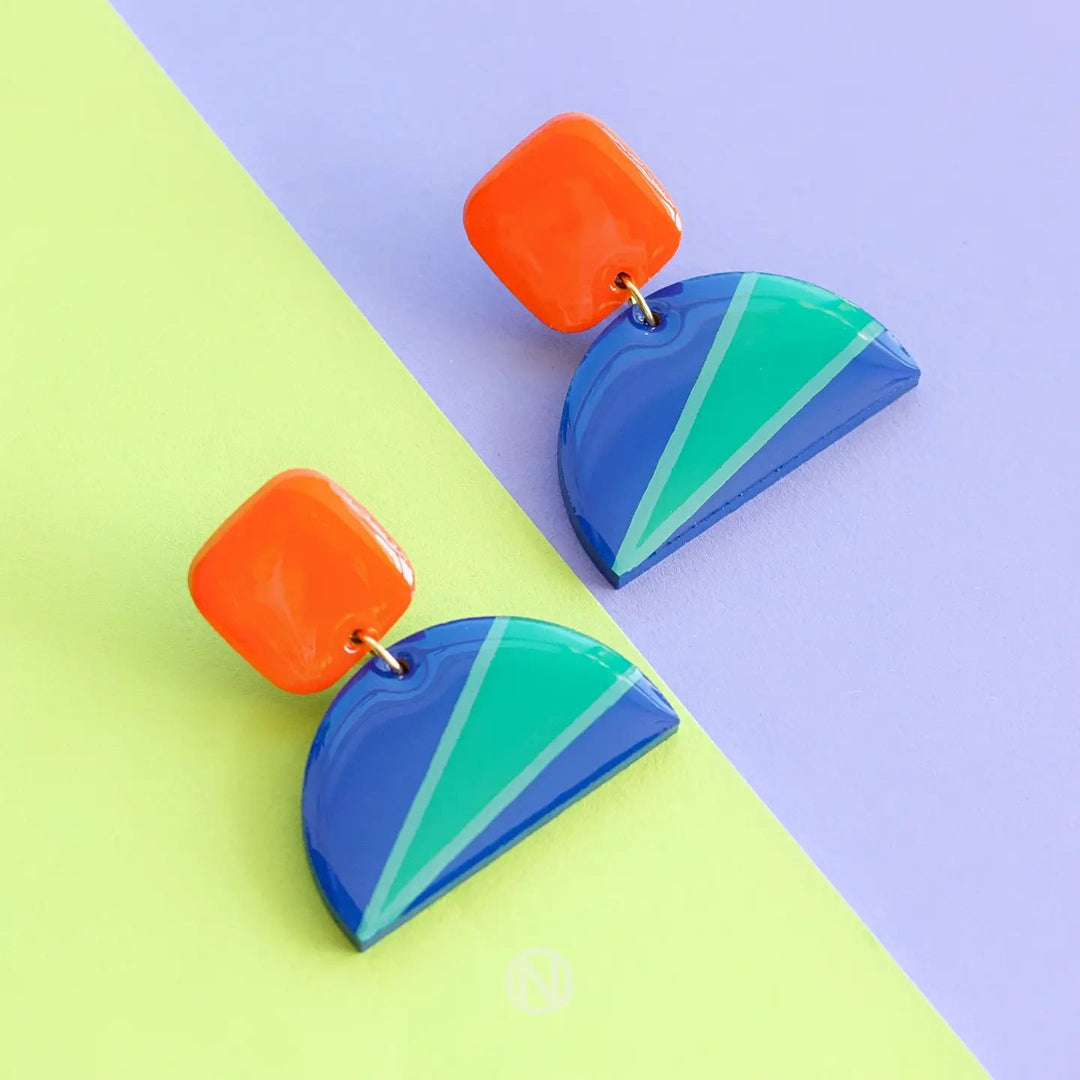 A pair of Vivid Statement Earrings by Naoi is displayed on a diagonal split background of yellow and purple. The wooden pendant earrings feature orange square tops connected to blue half-circle bottoms with green and teal triangular accents.