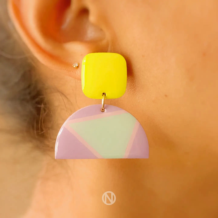 A close-up image of an ear wearing Vivid Statement Earrings by Naoi. The earring features a bright yellow square at the top and a semi-circle in light purple with a pastel green geometric pattern hanging below the square.