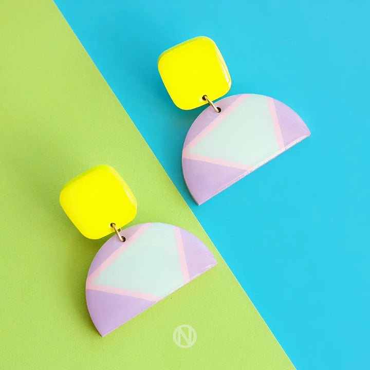 A pair of colorful, modern Naoi Vivid Statement Earrings is displayed against a divided background of green and blue. Each hand-painted earring features a bright yellow square stud connected to a semi-circle drop with a pastel purple, pink, and white geometric pattern.