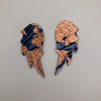 A pair of intricate Wings of Desire Sgraffito Stud Earrings by Rothlú with an abstract wing-like design. Sterling silver posts secure these artful pieces, which feature blue and black diagonal stripes interspersed with irregular gold patches, creating a striking contrast against the smooth, light background.