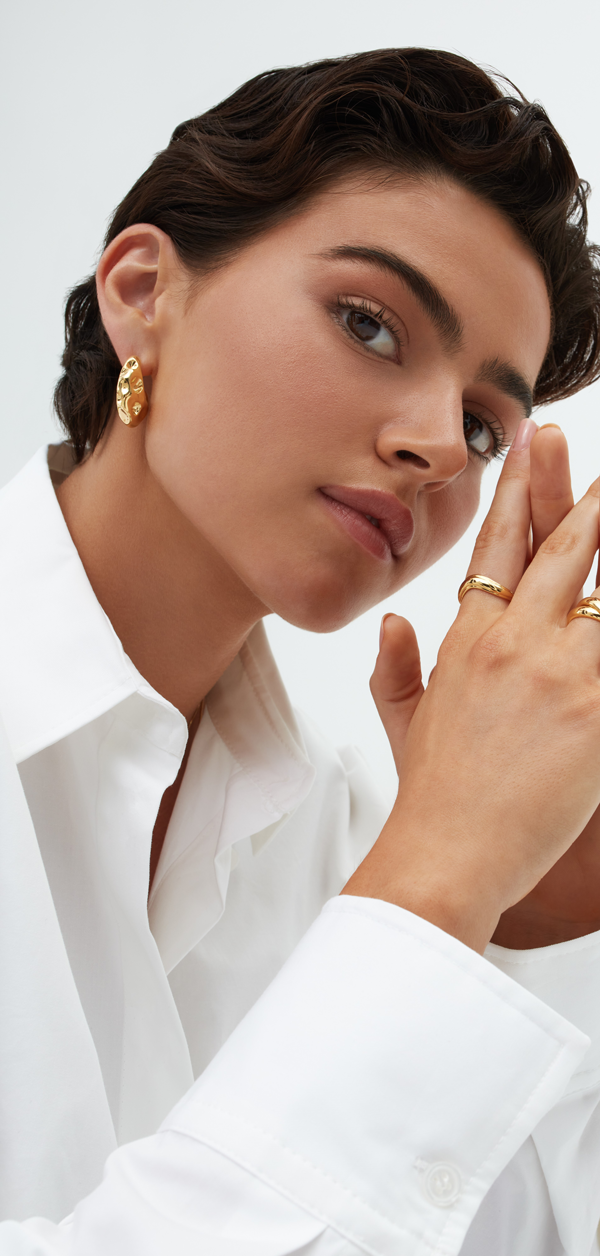 A person with short, dark hair and clear skin is wearing a white shirt and gold earrings shaped like crescents. They are looking directly at the camera with their hands near their face, displaying multiple Bonvo Wabi Rings with 18ct gold plating. The background is plain and light-colored.