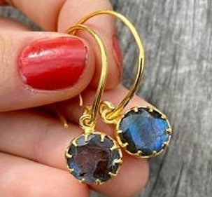 Close-up of a hand with red nail polish holding a pair of Labradorite Gold Dangles. The earrings, from Watermelon Tropical, feature round, iridescent labradorite gemstones set in a gold vermeil claw setting. The background is a textured gray surface, highlighting the beauty of this exquisite handmade jewellery.