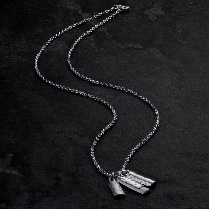 A silver necklace with a delicate chain is laid out on a dark, textured surface. The Lynsey de Burca Muir Cluster Pendant features three small, cylindrical pendants with subtle hammered detailing hanging from the chain.