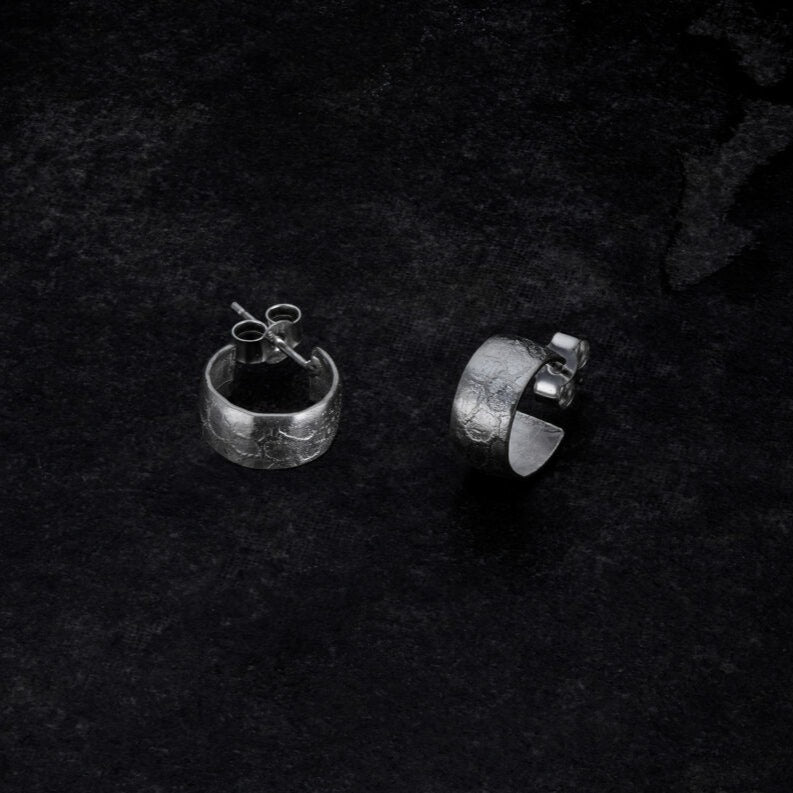 Two sterling silver earrings lie on a dark, textured surface. The Muir Sleepers from Lynsey de Burca boast a rough, hammered finish. One earring is lying flat with the post visible, while the other stands upright, showcasing its rounded shape and the artisanal touch of handmade jewellery.