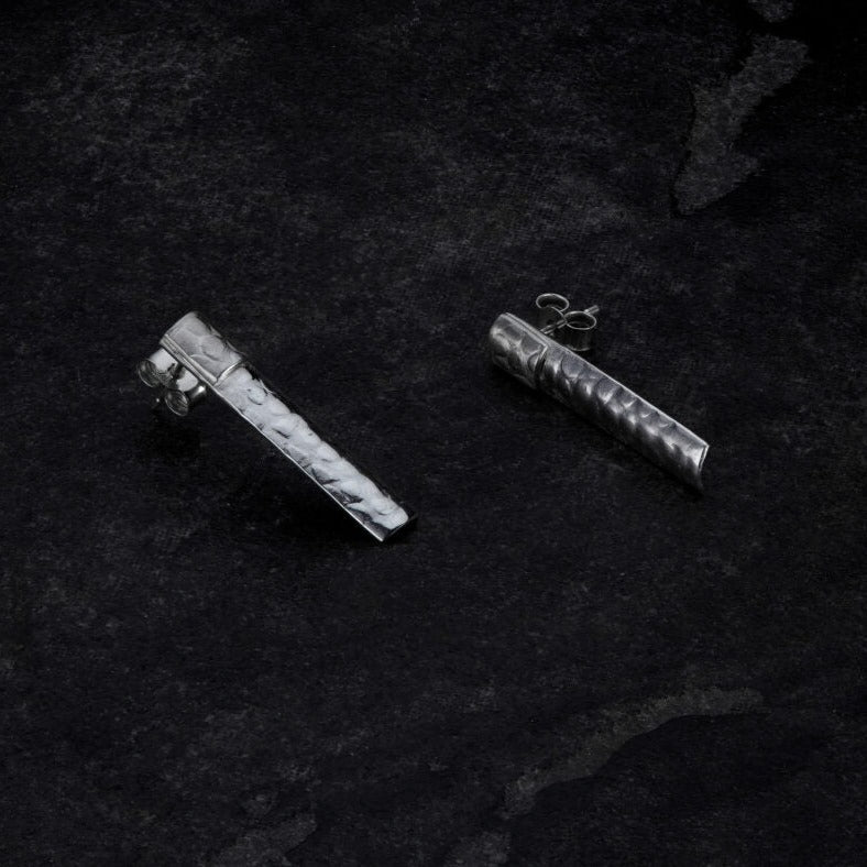 A pair of reflective, cylindrical, sterling silver Muir Studs with a textured pattern lay on a dark, rough surface. One earring rests on its side while the other stands upright, showcasing the exquisite handmade jewellery detailing by Lynsey de Burca.