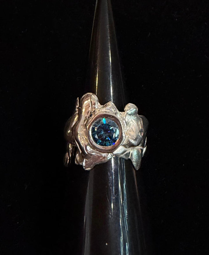 The Solstice Ring with Large Moissanite by Madeleine Sculptural Jewellery is a handcrafted silver ring with an organic, textured design, featuring a round moissanite center stone, displayed on a black glossy stand against a dark background.