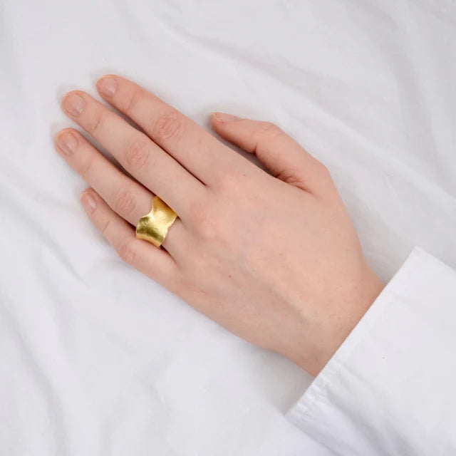 A hand wearing a crisp white long-sleeve shirt rests on a white surface, showcasing the Watermelon Tropical Wide Wavy Gold Plated Ring on the middle finger.