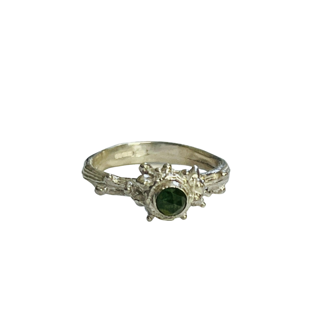 The Wild Woods Ring Green Tourmaline by Abbie Dixon is a silver twig ring featuring a dark green tourmaline gemstone surrounded by intricate, abstract detailing. The band has a textured appearance, complementing the unique design of the sterling silver ring. The overall look is vintage and elegant.