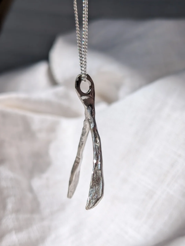 A close-up of a silver necklace with a textured, organic wish bone shaped pendant hanging from a silver curb chain against a soft, white fabric background.