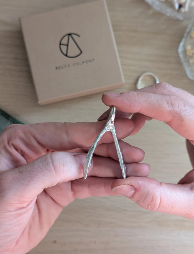 A person holds a Wishbone Pendant, crafted from recycled sterling silver. A beige box labeled “Becca Calmont” with a circular logo sits in the background on a wooden surface.