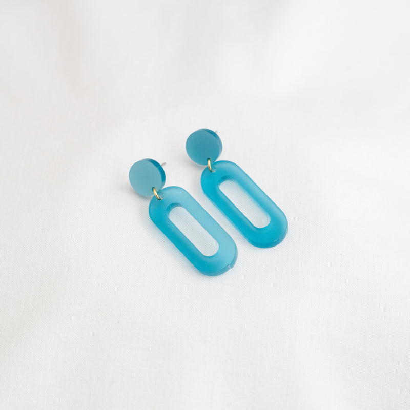 A pair of Timeless 04 Oval Acrylic Earrings by Studio Nok Nok featuring solid circular studs connected to elongated, hollow oval shapes, made from recycled materials, laid on a soft white fabric background.