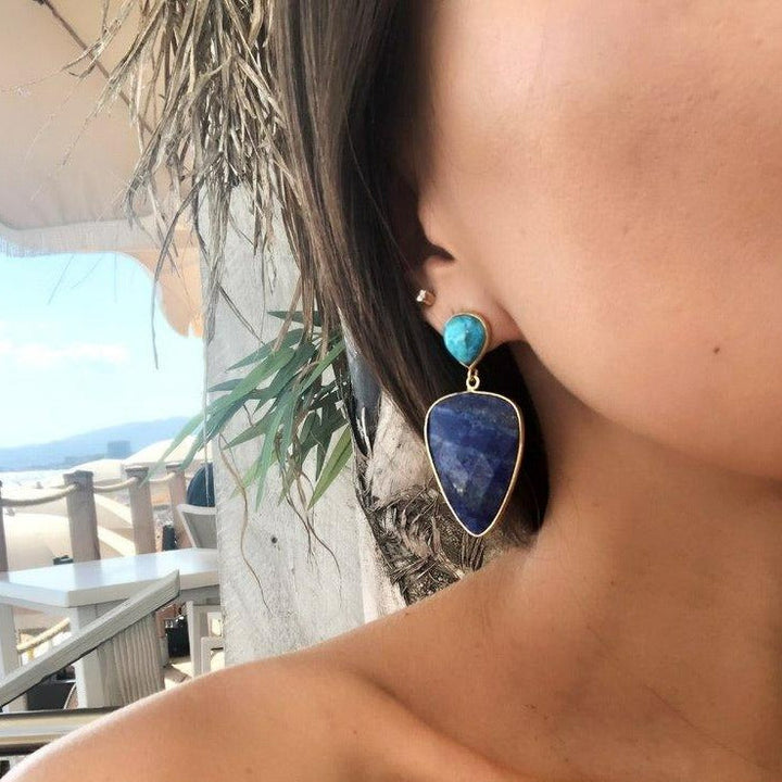 Close-up of a person’s ear adorned with Abi Earrings by Aria V. The earrings feature a small round turquoise stone at the top and a larger blue triangular lapis lazuli stone hanging below. In the background, an outdoor scene with part of a palm tree, a white umbrella, and clear sky is visible.